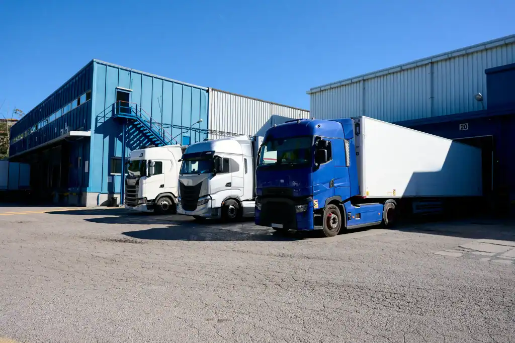 Three large trucks, two white and one blue, are parked at loading docks outside a blue industrial warehouse on a clear, sunny day, ready for Clean Truck Check CARB Compliance in Los Angeles & Riverside County, CA.