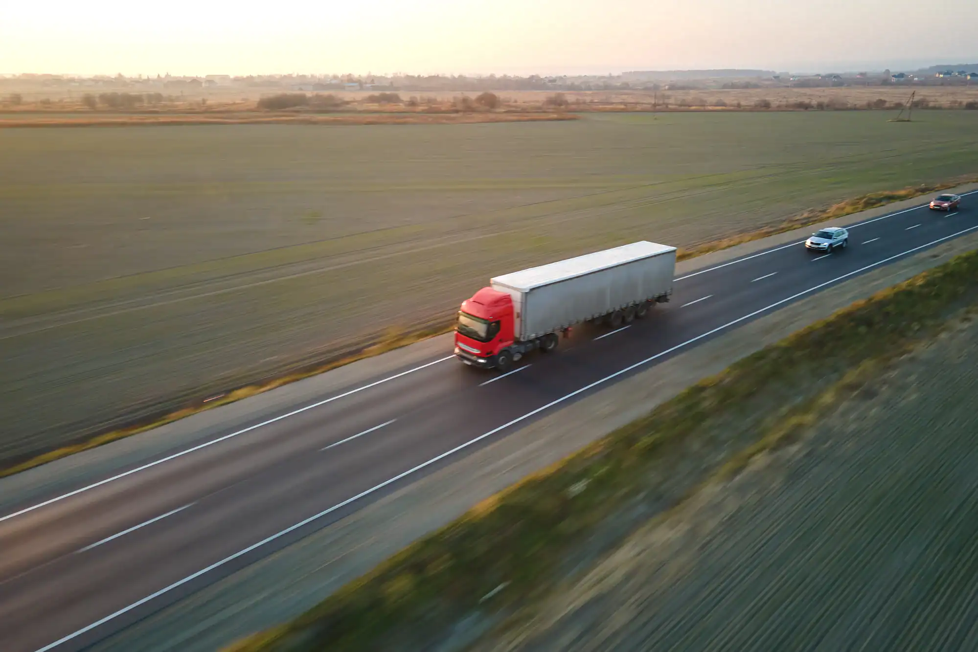 A red semi-truck with a gray trailer drives along a rural highway at sunset in Riverside County, carb Compliance required, as two cars travel the opposite way and open fields stretch on both sides of the road.