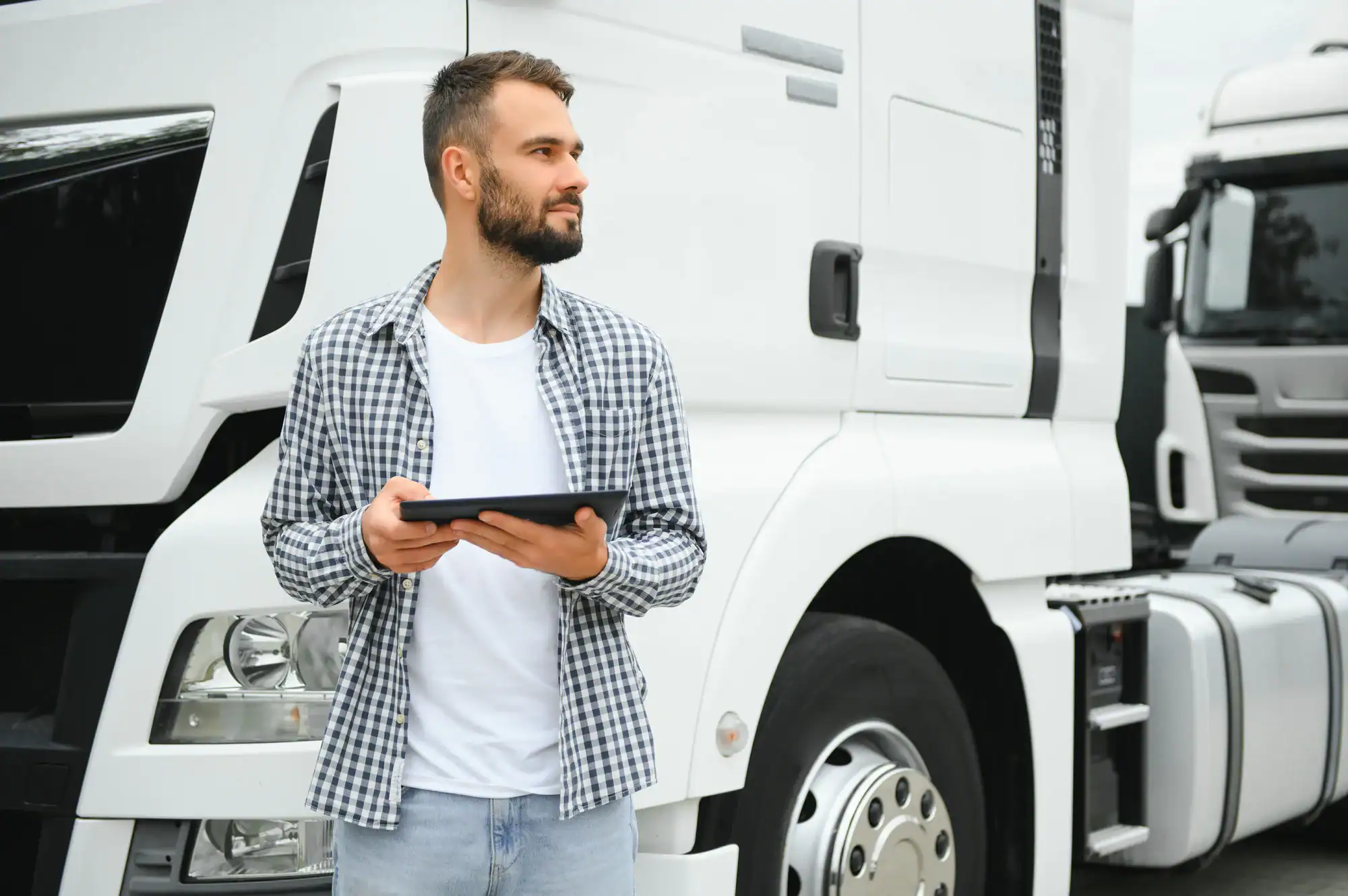 A man with a beard wearing a checkered shirt holds a tablet and stands in front of a large white truck in Los Angeles, ready for a Clean Truck Check or carb Compliance inspection. Another truck is visible in the background.