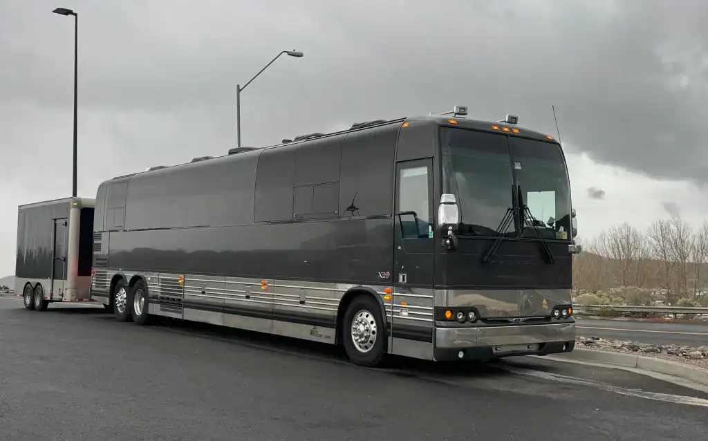 A large, dark gray tour bus with tinted windows is parked on a road under a cloudy sky, attached to a matching trailer.