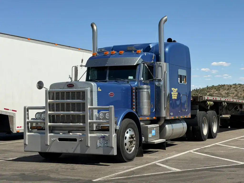 A blue semi-truck with a sleeper cab is parked in a lot next to a flatbed trailer and another truck on a sunny day, with clear skies and some trees in the background.