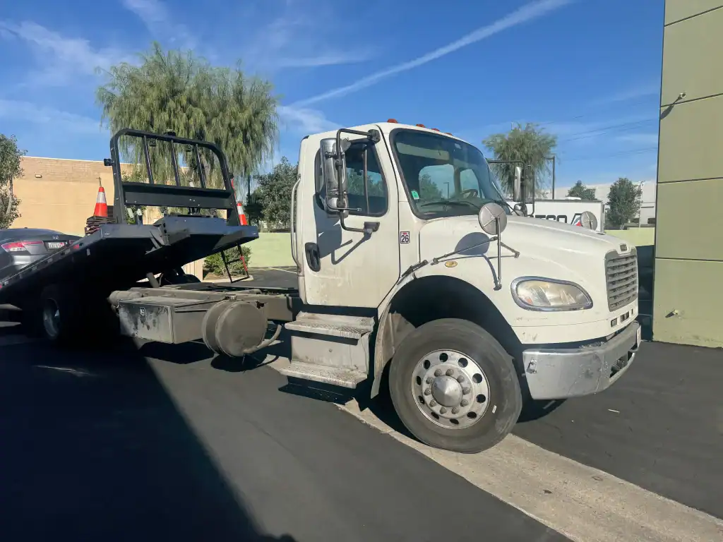 A white flatbed tow truck with its bed tilted down is parked on a sunny day near a green building and some trees.