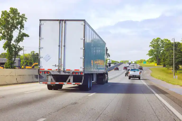 A large white semi-truck drives on a multi-lane highway in CA, alongside several cars, under partly cloudy skies with trees and grassy areas&mdash;highlighting the importance of Clean Truck Check Los Angeles & Riverside County initiatives.