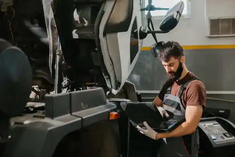 A mechanic wearing gloves and overalls uses a diagnostic laptop to inspect a large truck with its hood open inside a garage, performing a Clean Truck Check in Los Angeles & Riverside County, CA.