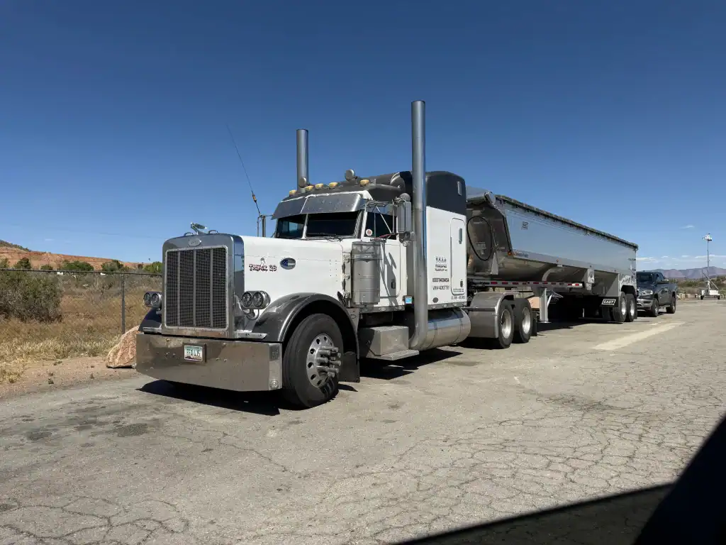 A large white semi-truck with a silver trailer is parked on cracked pavement under a clear blue CA sky, fenced in with dry vegetation—typical scenery for a Clean Truck Check in Los Angeles & Riverside County.