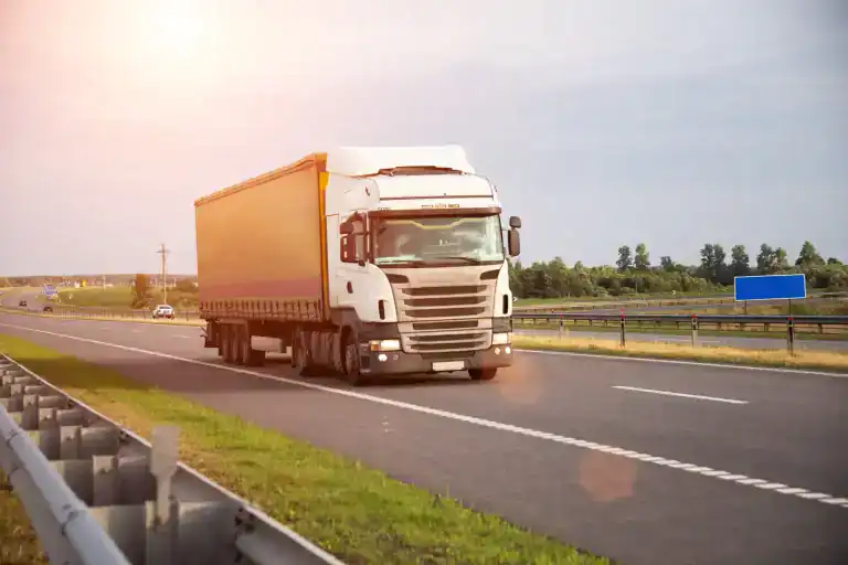 A large white semi-truck with a covered trailer, compliant with Clean Truck Check Los Angeles & Riverside County standards, drives on a CA highway in daylight, surrounded by green grass and trees under a clear sky.