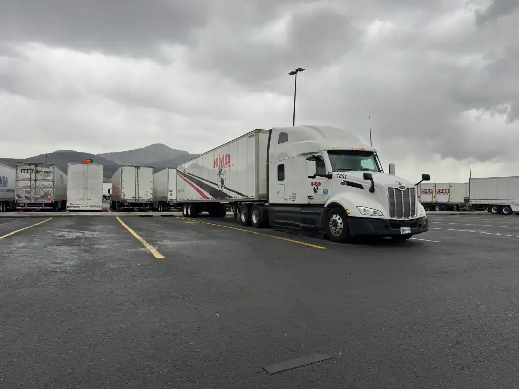 A white semi-truck with "HMD" on its trailer is parked in a CA lot on a cloudy day, surrounded by other trucks and distant mountains—ideal for Clean Truck Check Los Angeles & Riverside County compliance.