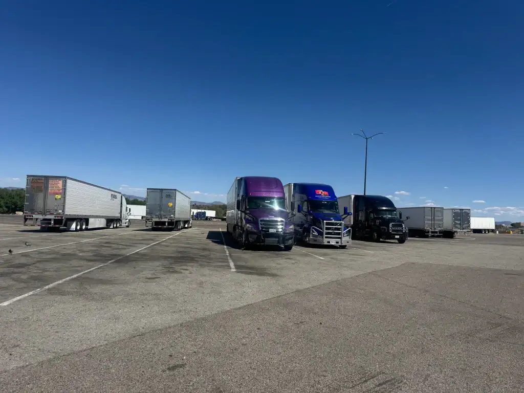 Several semi-trucks and trailers are parked in a mostly empty CA parking lot under a clear blue sky. The trucks, lined up side by side, hint at Clean Truck Check Los Angeles & Riverside County with mountains visible in the background.
