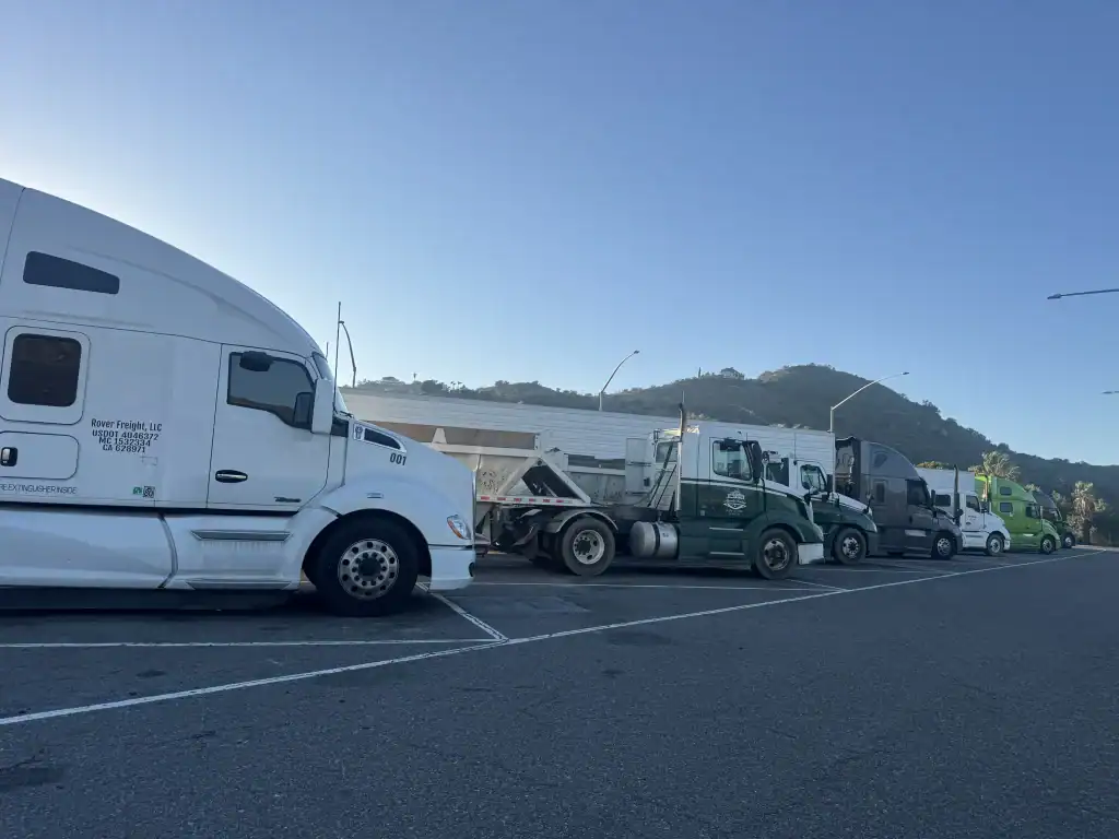 Several semi-trucks and trailers are parked in a row at a rest stop, with a hill and clear blue sky in the background. Mobile Clean Truck Check in LA & Riverside helps keep these rigs road-ready. The scene is calm and sunlit.