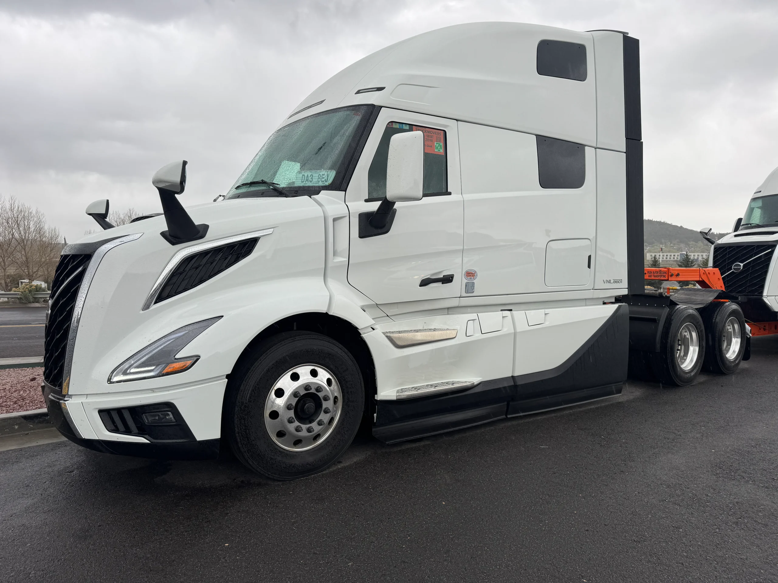 A white semi-truck with a modern design is parked on a wet road under a cloudy sky. The trailer is not attached, revealing the rear wheels and chassis. Another truck is partially visible in the background.